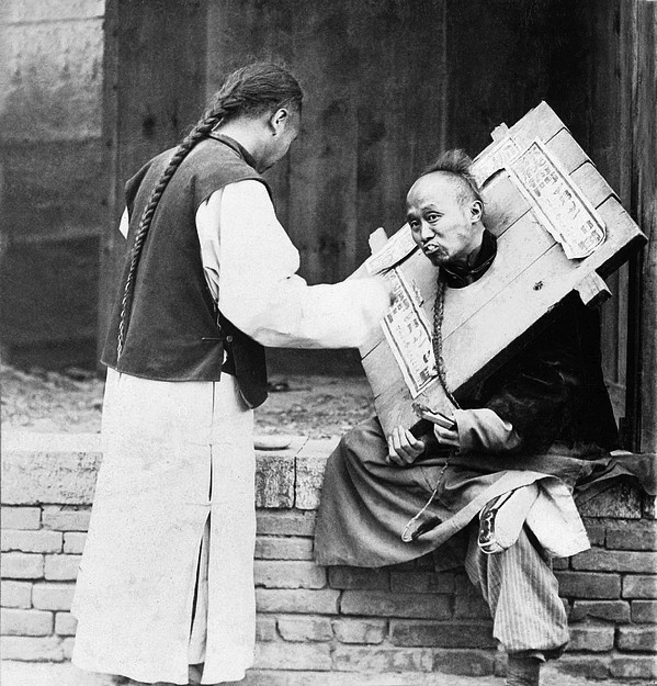 Chinese man feeding a criminal in a cangue with a sign describing his crime, 1905. Via